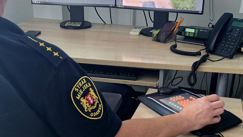 Na zdjęciu jest widoczna prawa ręka strażnika miejskiego w mundurze z logo Straż Miejska Gdańsk. Rękę trzyma na myszy komputerowej. Na biurku widoczny jest także czarny telefon stacjonarny i dolne fragmenty dwóch monitorów. 