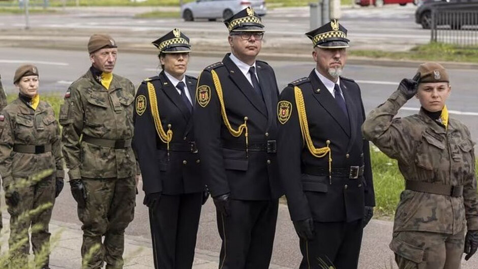 W szeregu stoją żołnierze i strażnicy miejscy. Pierwsza w szeregu żołnierka salutuje, za nią na baczności stoi troje strażników miejskich, a za nimi dwoje żołnierzy. 