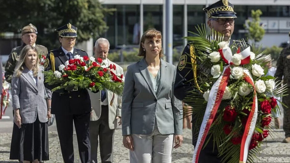 Strażnik miejski niesie wieniec biało-czerwonych kwiatów, za nim zastępca prezydent Gdańska Monika Chabior, za nią strażniczka z wieńcem kwiatów oraz kolejna oficjalna delegacja. 