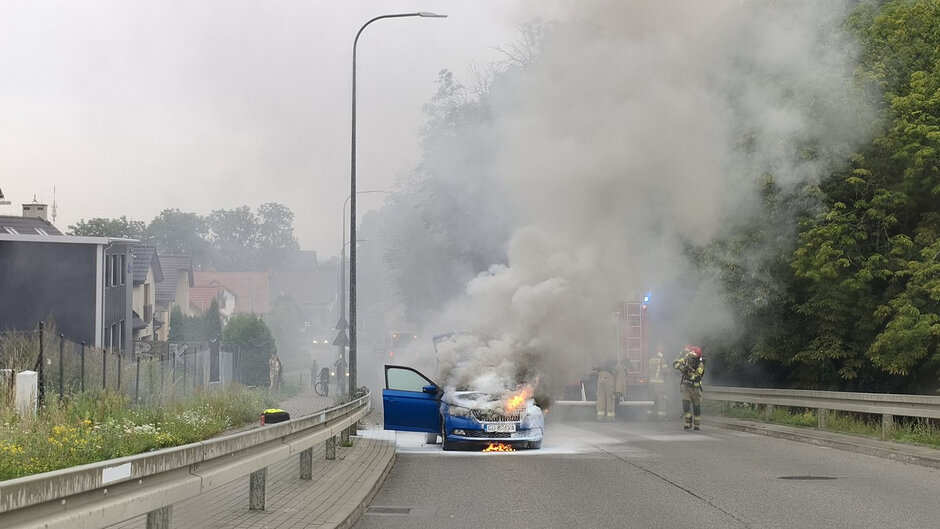 Na zdjęciu widoczny jest samochód osobowy koloru niebieskiego, który uległ pożarowi na jezdni. Ogień obejmuje przednią część pojazdu, szczególnie komorę silnika, skąd wydobywają się płomienie oraz gęste kłęby szaro-białego dymu unoszącego się nad drogą. Drzwi kierowcy są otwarte.
W pobliżu pojazdu prowadzone są działania ratowniczo-gaśnicze. W tle znajduje się samochód pożarniczy z włączonymi sygnałami świetlnymi, a obok niego strażacy wyposażeni w ubrania specjalne i sprzęt ochrony osobistej. Na jezdni widoczne są ślady użycia środków gaśniczych.
Zdarzenie ma miejsce na drodze przebiegającej pomiędzy zabudową mieszkaniową a terenem zadrzewionym. Zdjęcie dokumentuje akcję gaśniczą prowadzoną w celu ugaszenia pożaru pojazdu i zabezpieczenia miejsca zdarzenia.