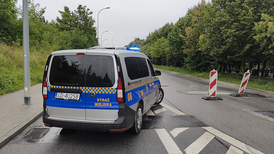 Na fotografii przedstawiono oznakowany radiowóz Straży Miejskiej Gdańska marki Volkswagen, ustawiony na jezdni z włączonym niebieskim sygnałem świetlnym. Pojazd częściowo blokuje ruch drogowy w rejonie prowadzonej interwencji.
Przed radiowozem ustawiono przenośne zapory drogowe w czerwono-białe pasy, wyznaczające strefę wyłączoną z ruchu. Nawierzchnia jezdni jest mokra, co może być związane z prowadzonymi wcześniej działaniami gaśniczymi.
Po obu stronach drogi znajdują się tereny zielone porośnięte drzewami i krzewami. Zdjęcie dokumentuje działania Straży Miejskiej polegające na zabezpieczeniu miejsca zdarzenia oraz kierowaniu ruchem w czasie prowadzenia akcji ratowniczo-gaśniczej.
