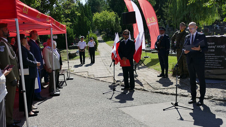 Na Cmentarzu Łostowickim, przy pomniku upamiętniającym ofiary “operacji polskiej”, przemawia dyrektor Piotr Kryszewski. Obok stoi mężczyzna prowadzący uroczystość, a naprzeciwko - jej uczestnicy. 