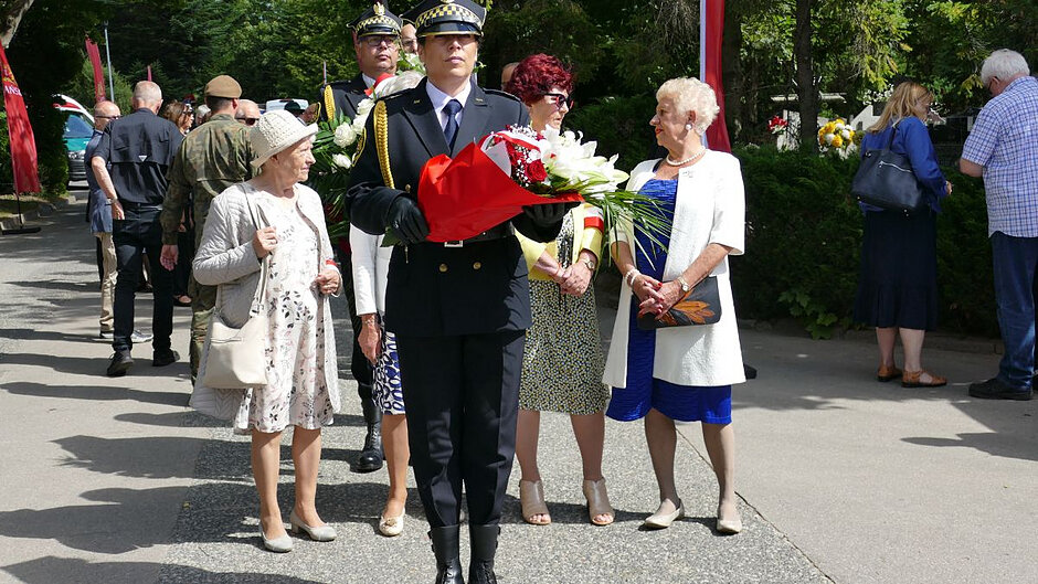 Strażniczka miejska w galowym mundurze stoi z wiązanką kwiatów, za nią delegacja składająca się z trzech kobiet, a następnie kolejny strażnik miejski. 