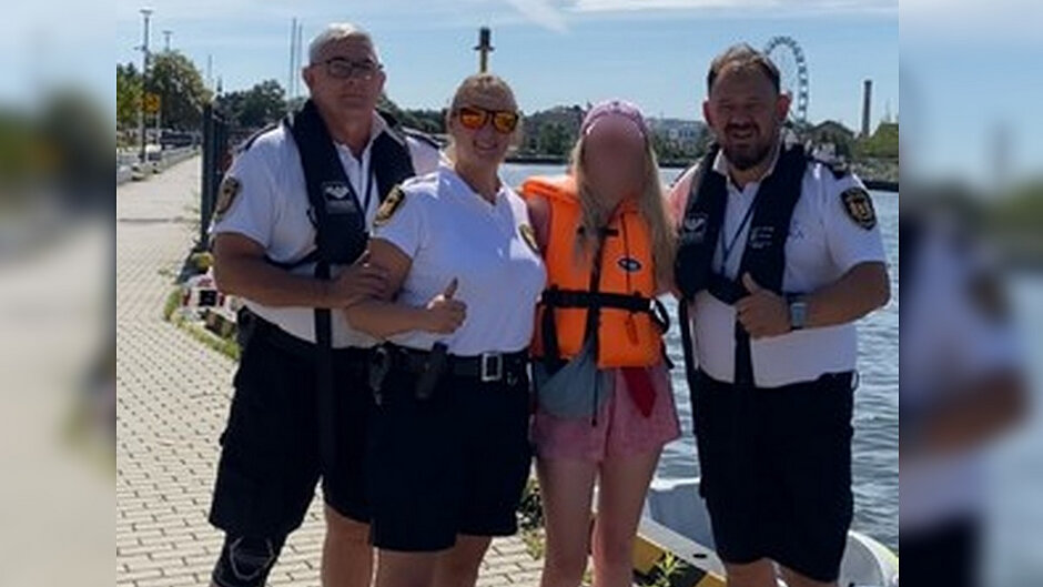 Na nabrzeżu nad wodą wykonano pamiątkowe zdjęcie grupowe. Cztery osoby stoją obok siebie, a jedna z nich ma na sobie kamizelkę ratunkową. W tle widoczny jest akwen wodny, elementy infrastruktury portowej oraz koło widokowe. Zdjęcie dokumentuje wydarzenie związane z promocją bezpieczeństwa nad wodą i współpracą służb z uczestnikami akcji.