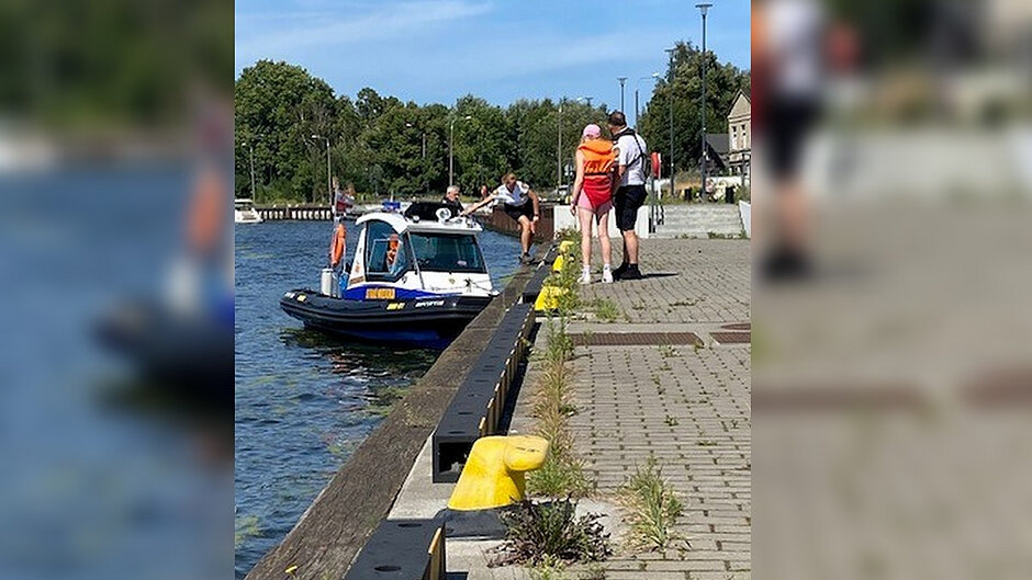 Przy nabrzeżu zacumowana jest łódź patrolowa straży miejskiej. Na pomoście znajdują się osoby uczestniczące w działaniach związanych z bezpieczeństwem na wodzie. Zdjęcie przedstawia fragment nabrzeża, akwen wodny oraz jednostkę pływającą wykorzystywaną do patroli i działań ratowniczych.