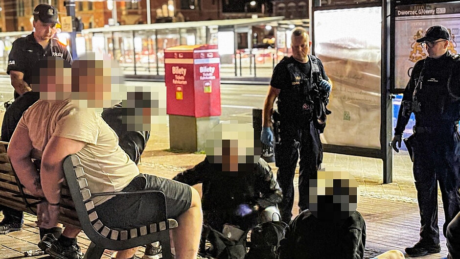 Na zdjęciu przedstawiono interwencję służb porządkowych prowadzoną w porze nocnej w rejonie przystanku komunikacji miejskiej. W centralnej części kadru znajduje się kilka osób siedzących na ławce oraz na nawierzchni peronu. Twarze osób zostały zamazane. Wokół zgromadzonych osób znajdują się funkcjonariusze Straży Miejskiej ubrani w czarne umundurowanie służbowe. Jeden z funkcjonariuszy wykonuje czynności bezpośrednio przy osobach siedzących na ziemi, natomiast pozostali zabezpieczają miejsce zdarzenia i obserwują przebieg interwencji.

W tle widoczna jest infrastruktura przystankowa oraz tablica informacyjna. Na znajdującym się obok przystanku słupku można odczytać nazwę „Dworzec Główny”. Widoczne są również dwa czerwone pojemniki oznaczone napisem „Bilety”. Dalszy plan tworzą elementy infrastruktury komunikacyjnej oraz oświetlone budynki miejskie.
Zdjęcie wykonano po zmroku przy sztucznym oświetleniu ulicznym i przystankowym. Fotografia dokumentuje działania interwencyjne Straży Miejskiej prowadzone w przestrzeni publicznej w rejonie Dworca Głównego w Gdańsku.