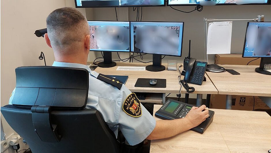 Tyłem siedzi operator monitoringu, a przed nim trzy ekrany komputerowe z rozmazanym zapisem z kamer. Na stole leży telefon i krótkofalówka.