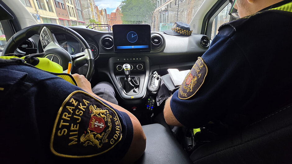 Na zdjęciu przedstawiono wnętrze oznakowanego pojazdu służbowego Straży Miejskiej w Gdańsku podczas patrolu miejskiego. Fotografia została wykonana z tylnej części samochodu i ukazuje dwóch funkcjonariuszy siedzących na przednich fotelach. Na rękawach mundurów widoczne są naszywki z napisem „Straż Miejska” oraz herbem Gdańska. W centralnej części kabiny znajduje się ekran multimedialny wyświetlający symbol zegara, a poniżej panel sterowania i dźwignia manualnej skrzyni biegów. Między siedzeniami widoczny jest radiotelefon oraz notatnik wykorzystywany podczas służby. Na desce rozdzielczej leży czapka służbowa. Przez przednią szybę można zauważyć zabudowę śródmiejską z kamienicami oraz historycznym budynkiem w tle. Zdjęcie dokumentuje codzienną pracę patrolu Straży Miejskiej podczas wykonywania obowiązków służbowych na terenie miasta.