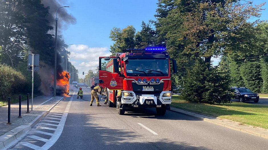 Na miejscu pożaru prowadzone są działania gaśnicze. Na pierwszym planie znajduje się wóz Państwowej Straży Pożarnej z włączonymi sygnałami świetlnymi. W tle strażacy przygotowują sprzęt do gaszenia płonącego samochodu dostawczego, nad którym nadal unoszą się kłęby czarnego dymu. Miejsce zdarzenia zostało zabezpieczone na czas akcji ratowniczej.