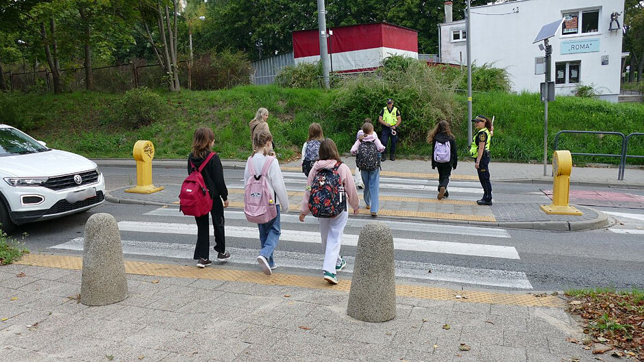 Kilkoro dzieci z plecakami na plecach przechodzi przez przejście dla pieszych. Pośrodku stoi strażniczka miejska, po drugiej stronie ulicy stoi strażnik miejski. Obydwoje pomagają dzieciom przejść na drugą stronę.