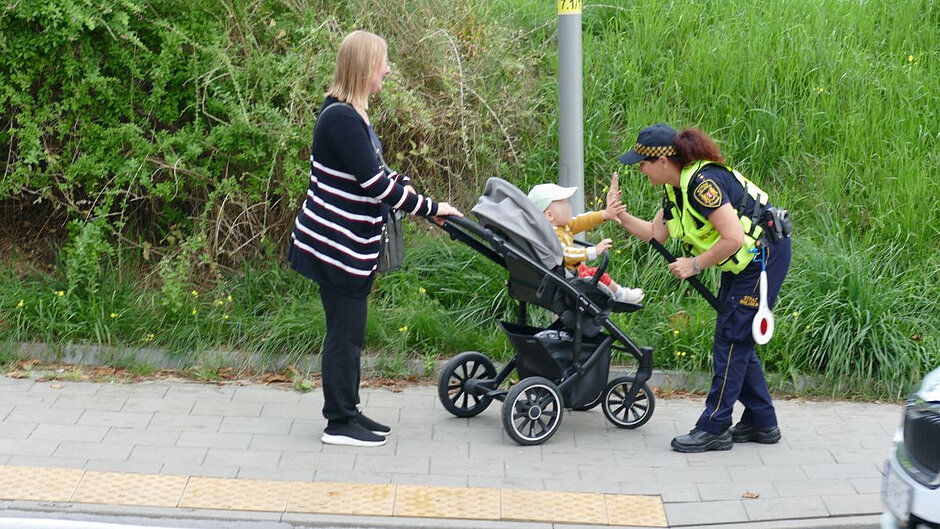 Na chodniku stoi kobieta z wózkiem, w którym siedzi małe dziecko. Maluch przybija piątkę ze strażniczką miejską.
