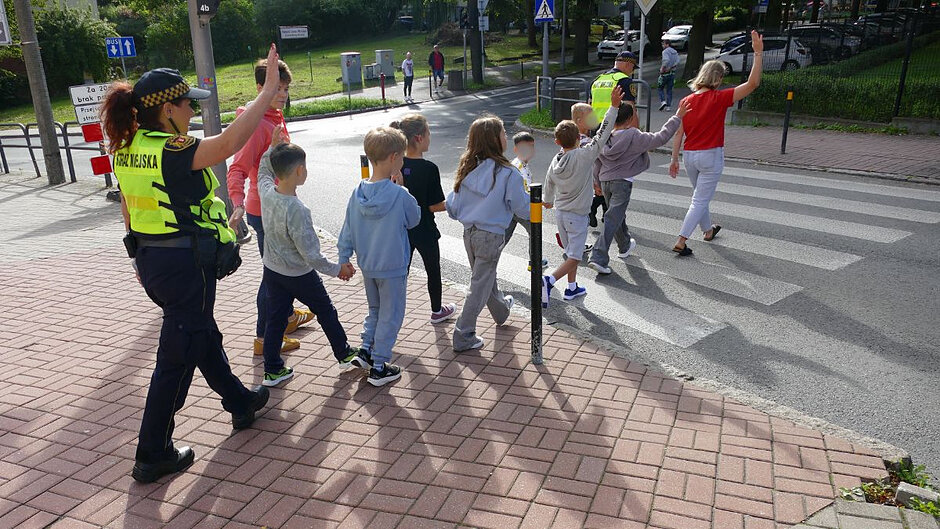 Dzieci w parach przechodzą przez przejście dla pieszych. Towarzyszą im dwie nauczycielki oraz dwójka strażników miejskich.