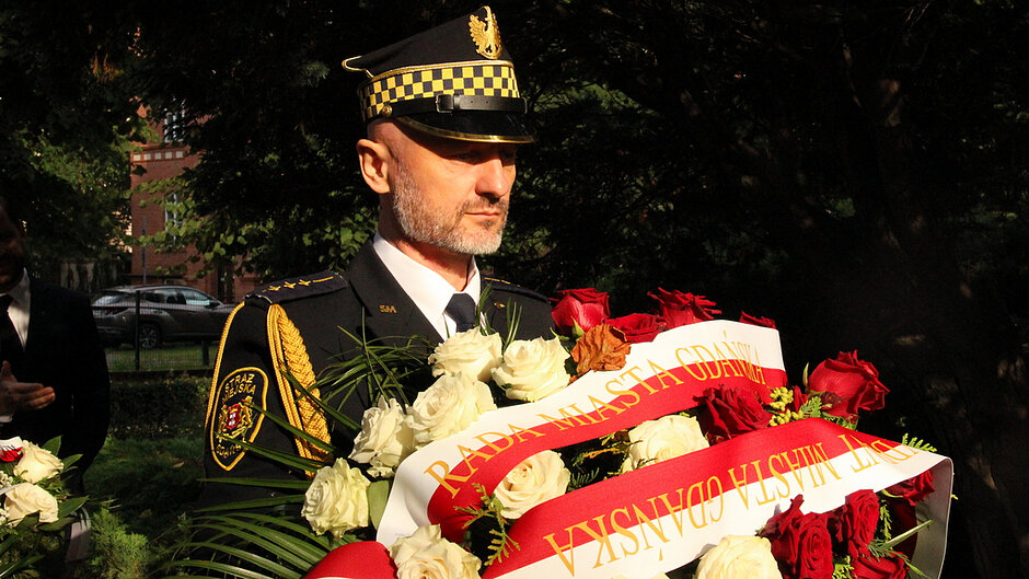 Na zdjęciu widoczna jest osoba w umundurowaniu Straży Miejskiej trzymająca okazałą wiązankę czerwonych i białych róż przewiązaną biało-czerwonymi szarfami. Na szarfach znajduje się napis „Rada Miasta Gdańska”. Fotografia została wykonana w plenerze, w otoczeniu drzew i zieleni. Kwiaty stanowią symbol pamięci i hołdu składanego podczas uroczystości o charakterze patriotycznym lub rocznicowym.
