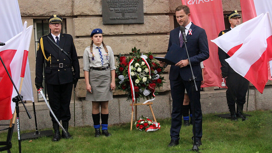 Na zdjęciu uwieczniono przemówienie wygłaszane podczas uroczystości upamiętniającej. Przed tablicą pamiątkową ustawiono wieniec z biało-czerwonych kwiatów, przy którym wartę pełnią harcerze. W ceremonii uczestniczą również przedstawiciele Straży Miejskiej. Widoczne są liczne flagi państwowe oraz elementy oprawy oficjalnej, podkreślające patriotyczny charakter wydarzenia.