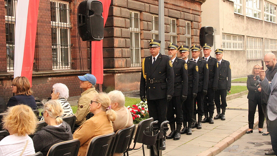 Poczet honorowy Straży Miejskiej uczestniczy w obchodach rocznicowych organizowanych na terenie miasta. Funkcjonariusze ustawieni w szyku stoją w pobliżu miejsca składania kwiatów, obok zgromadzonych uczestników uroczystości. Wydarzenie ma podniosły charakter i stanowi wyraz szacunku dla pamięci historycznej oraz lokalnego dziedzictwa.