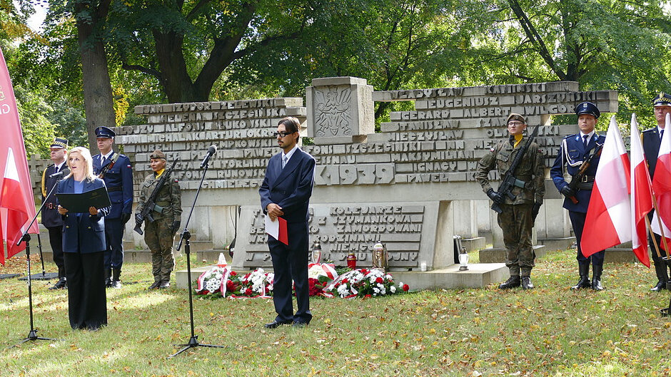 Przemówienie podczas uroczystości upamiętniającej ofiary II wojny światowej przy pomniku zawierającym nazwiska poległych. W ceremonii uczestniczą przedstawiciele władz, poczty sztandarowe oraz żołnierze pełniący wartę honorową. U stóp pomnika znajdują się złożone wiązanki kwiatów i znicze, stanowiące wyraz pamięci i szacunku dla bohaterów.