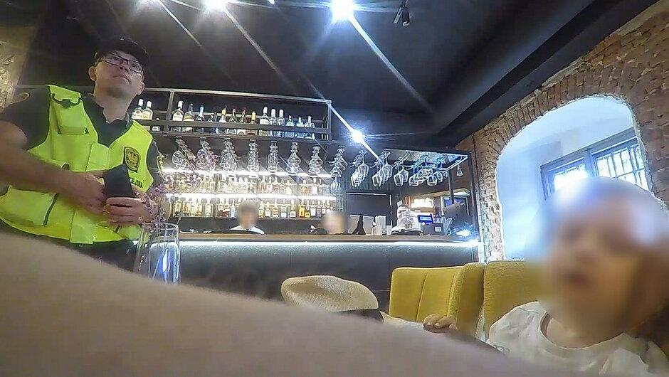 Zdjęcie wykonane we wnętrzu lokalu gastronomicznego. Po lewej stronie kadru widoczna jest osoba ubrana w jaskrawozieloną kamizelkę z emblematem oraz ciemne nakrycie głowy, trzymająca w dłoniach ciemny przedmiot przypominający telefon lub niewielkie urządzenie. W tle znajduje się bar z podświetlanym blatem, półkami wypełnionymi butelkami oraz zawieszonymi kieliszkami. Po prawej stronie kadru znajduje się dziecko siedzące na jasnym fotelu lub kanapie. Twarz dziecka jest zamazana. W dolnej części zdjęcia widoczny jest nieostry fragment obiektu zasłaniający część kadru. Wnętrze lokalu posiada ceglane ściany, duże okno oraz oświetlenie sufitowe.