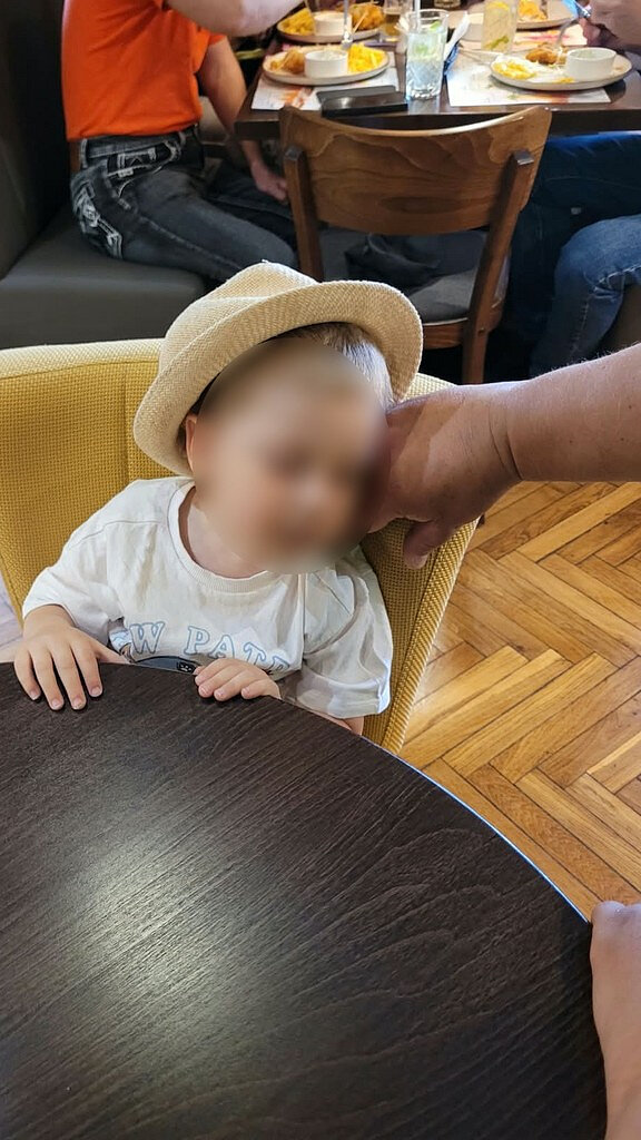 Zdjęcie przedstawia dziecko siedzące przy ciemnym, okrągłym stole w lokalu gastronomicznym. Dziecko ma na głowie jasny kapelusz, a twarz została zamazana. Obok głowy dziecka znajduje się dłoń osoby dorosłej. W tle widoczny jest stół, przy którym siedzą inne osoby, talerze z jedzeniem, szklanki oraz telefon komórkowy. Na podłodze znajduje się drewniany parkiet ułożony w jodełkę. Meble mają jasne i ciemne odcienie drewna oraz tapicerowane siedziska w kolorze żółtym i szarym. Na koszulce dziecka częściowo widoczny jest napis rozpoczynający się od liter „NEW”.