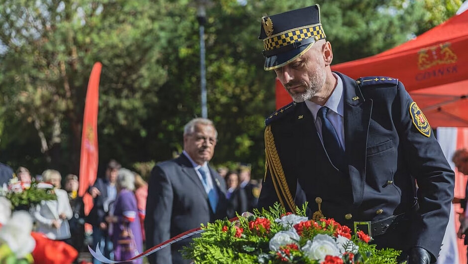 Drugie zdjęcie przedstawia moment niesienia wieńca przez przedstawiciela Straży Miejskiej w Gdańsku. W tle widoczni są reprezentanci innych służb mundurowych oraz pozostali uczestnicy uroczystości. Biało-czerwone szarfy i kwiaty na wieńcu nawiązują do narodowego charakteru obchodów. Zdjęcie ukazuje współudział różnych formacji i instytucji w pielęgnowaniu pamięci historycznej.