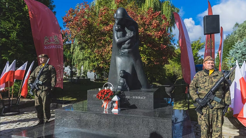 Na trzecim zdjęciu widoczny jest Pomnik Ofiar Golgoty Wschodu, przed którym złożono wiązankę kwiatów. Po obu stronach monumentu wartę honorową pełnią żołnierze Wojska Polskiego. Wokół pomnika znajdują się biało-czerwone flagi oraz miejskie flagi Gdańska. Słoneczna pogoda i otaczająca zieleń tworzą godną oprawę dla uroczystości poświęconej pamięci osób deportowanych na Sybir i ofiar represji totalitarnych.