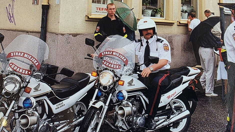 Zdjęcie z lat 90. ubiegłego wieku. Widać na nim dwa motocykle Straży Miejskiej w Gdańsku. Na jednym z nich siedzi strażnik. Za nim stoi inny strażnik.