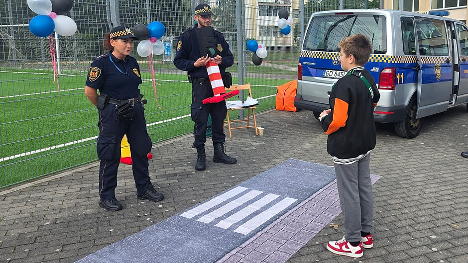 Trzecia fotografia przedstawia praktyczne zajęcia edukacyjne z wykorzystaniem miasteczka ruchu drogowego. Uczestnik ćwiczy prawidłowe przechodzenie przez przejście dla pieszych, a strażnicy wyjaśniają najważniejsze zasady bezpieczeństwa.