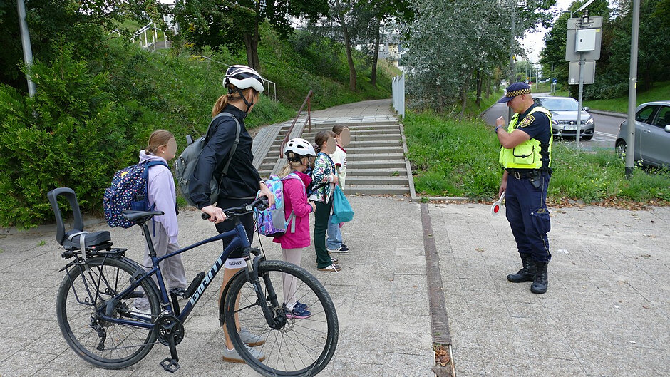 widać zajęcia edukacyjne prowadzone przez strażnika miejskiego dla grupy dzieci w wieku szkolnym. Spotkanie odbywa się w pobliżu ciągu pieszego i schodów prowadzących do osiedlowej drogi. Uczestnicy słuchają instrukcji dotyczących zasad bezpiecznego poruszania się po drogach i zachowania ostrożności podczas codziennych dojść do szkoły. Obok dzieci znajduje się rower, który stanowi dodatkowy element zajęć związanych z bezpieczeństwem niechronionych uczestników ruchu drogowego. W tle widoczne są drzewa, chodnik oraz ruch uliczny, co pozwala prowadzić naukę w warunkach zbliżonych do rzeczywistych sytuacji spotykanych na co dzień.