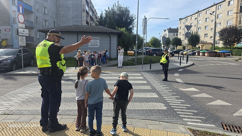 Na pierwszym zdjęciu strażnicy miejscy pomagają grupie najmłodszych bezpiecznie przejść przez przejście dla pieszych. Dzieci pod opieką funkcjonariuszy ćwiczą prawidłowe zachowania na drodze w rzeczywistych warunkach miejskich.
