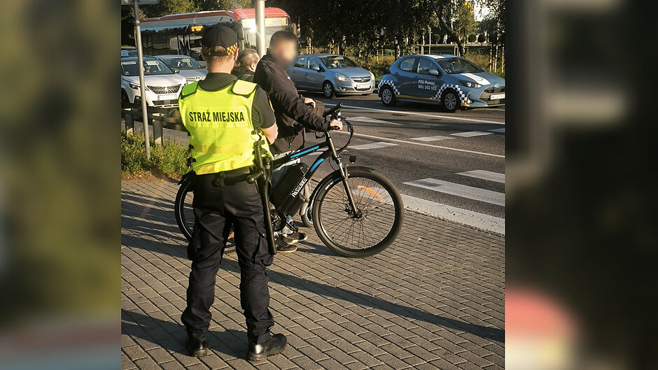 Na drugim zdjęciu funkcjonariusz Straży Miejskiej prowadzi rozmowę z rowerzystą, który zatrzymał się przy przejściu dla pieszych i przejeździe rowerowym. W tle widoczny jest ruch uliczny oraz oznakowany pojazd pomocy drogowej. Fotografia przedstawia jedną z kontroli prowadzonych wobec użytkowników jednośladów, mających na celu przypominanie zasad bezpiecznego poruszania się po drogach, ścieżkach rowerowych i przejazdach dla rowerów. Kadr pokazuje codzienną służbę strażników miejskich oraz działania profilaktyczne realizowane na terenie miasta.