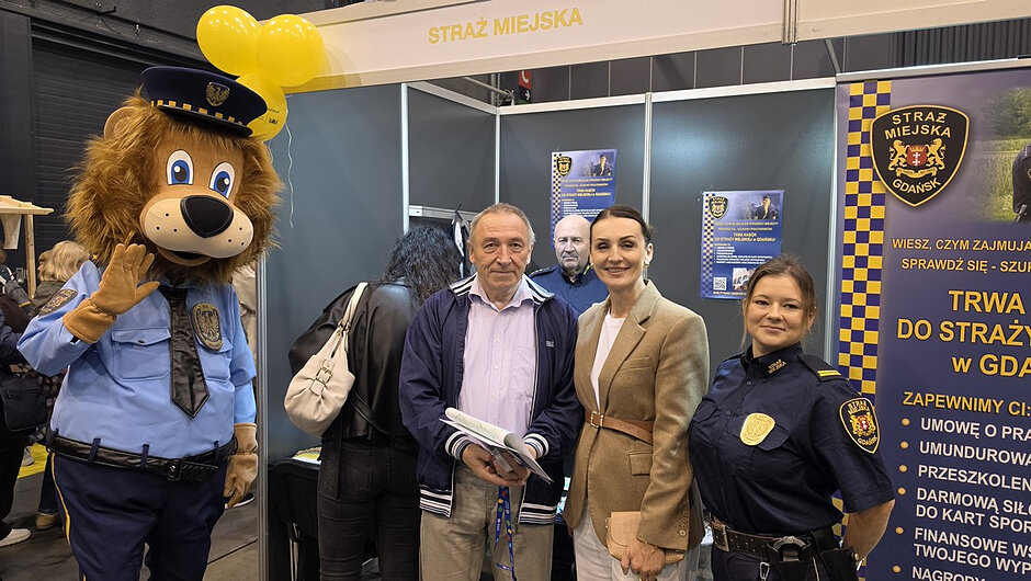 Przy stoisku Straży Miejskiej w Gdańsku znajdującym się na terenie hali targowej pozuje kilka osób oraz maskotka formacji, lew Leoś. Po lewej stronie kadru stoi wysoka maskotka ubrana w błękitny mundur strażnika miejskiego i granatową czapkę. W centralnej części zdjęcia widać osoby odwiedzające stoisko oraz funkcjonariuszkę Straży Miejskiej. Nad stoiskiem umieszczono napis „Straż Miejska”, a w tle znajdują się materiały informacyjne, roll-up z logo formacji oraz żółte balony. Zdjęcie pokazuje bezpośredni kontakt strażników z mieszkańcami i dużą popularność stoiska podczas targów.