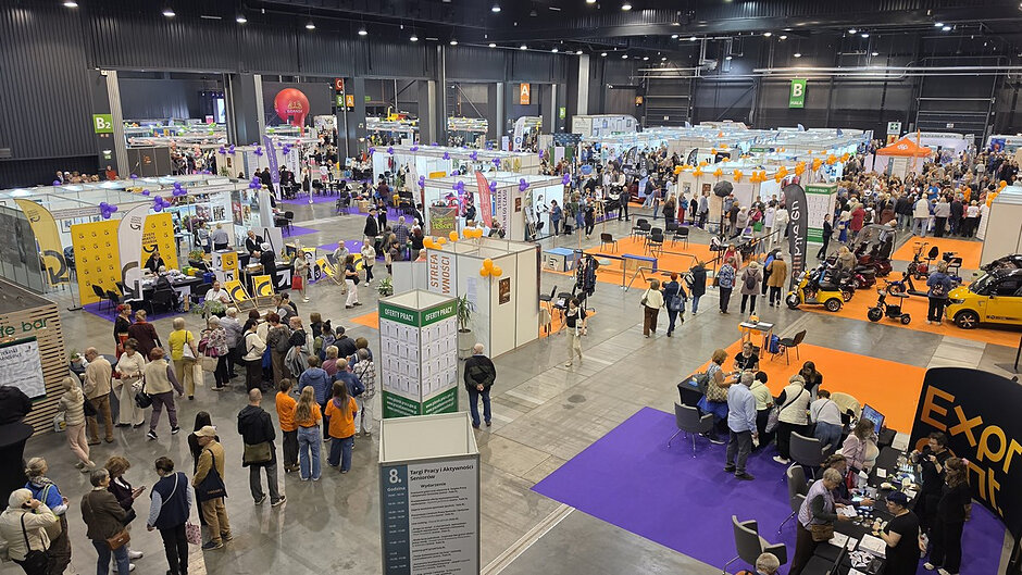 Widok ogólny na halę AmberExpo podczas VIII Targów Pracy i Aktywności Seniorów. Na dużej przestrzeni rozmieszczono dziesiątki stoisk wystawców reprezentujących instytucje publiczne, organizacje społeczne oraz pracodawców. Między stoiskami poruszają się liczni odwiedzający. Widoczne są strefy spotkań, miejsca konsultacji, punkty informacyjne oraz kolorowe dekoracje oznaczające poszczególne sektory wydarzenia. Zdjęcie ukazuje skalę targów oraz bardzo wysoką frekwencję uczestników.