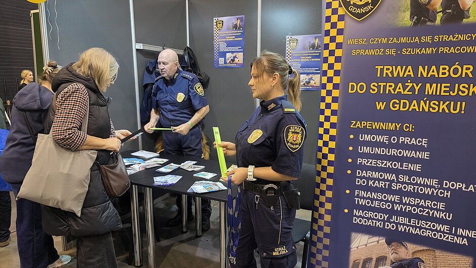 Stoisko Straży Miejskiej podczas VIII Targów Pracy i Aktywności Seniorów. Na pierwszym planie funkcjonariuszka rozmawia z uczestniczką wydarzenia i przekazuje materiały informacyjne. Za stolikiem widoczny jest drugi strażnik miejski, który odpowiada na pytania odwiedzających. Na stole znajdują się ulotki, broszury i gadżety profilaktyczne. Po prawej stronie kadru ustawiono duży baner informujący o naborze do Straży Miejskiej w Gdańsku. Zdjęcie dokumentuje działania edukacyjne i profilaktyczne prowadzone przez strażników podczas targów.