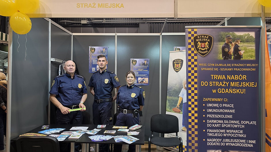 Stoisko Straży Miejskiej w Gdańsku podczas VIII Targów Pracy i Aktywności Seniorów w AmberExpo. Za stolikiem z materiałami informacyjnymi stoją troje umundurowanych strażników miejskich, którzy prezentują ofertę formacji i odpowiadają na pytania odwiedzających. Na stole rozłożone są ulotki, broszury oraz gadżety profilaktyczne. Po prawej stronie zdjęcia widoczny jest duży baner zachęcający do wstąpienia do Straży Miejskiej w Gdańsku, zawierający informacje o prowadzonym naborze oraz warunkach zatrudnienia. Nad stoiskiem znajduje się napis „Straż Miejska”, a jego przestrzeń ozdobiono żółtymi balonami. Zdjęcie przedstawia przygotowane stanowisko informacyjno-profilaktyczne gdańskich strażników podczas targów.