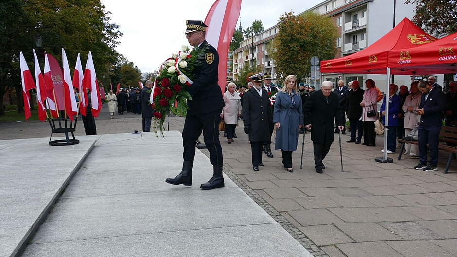 Strażnik miejski niesie wieniec reprezentacji miasta Gdańska i środowisk kombatanckich. W tle zaproszeni goście oraz biało-czerwone flagi. 
