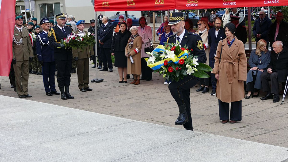 Strażnik miejski niesie wieniec, za nim konsul Ukrainy w Gdańsku, w tle uczestnicy wydarzenia oraz kolejny strażnik z kwiatami i delegacja związku kombatantów.