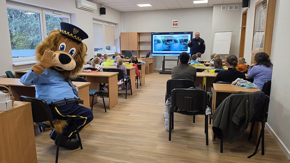 W sali szkoleniowej przy drewnianych ławkach siedzi grupa dzieci uczestniczących w zajęciach edukacyjnych. Na pierwszym planie widoczne jest dziecko trzymające w dłoniach okrągły odblask z napisem „1986 Straż Miejska”. Na oparciach krzeseł i ławek leżą jaskrawożółte kamizelki odblaskowe. Dzieci siedzą tyłem do obiektywu i skupiają uwagę na prowadzonych zajęciach.