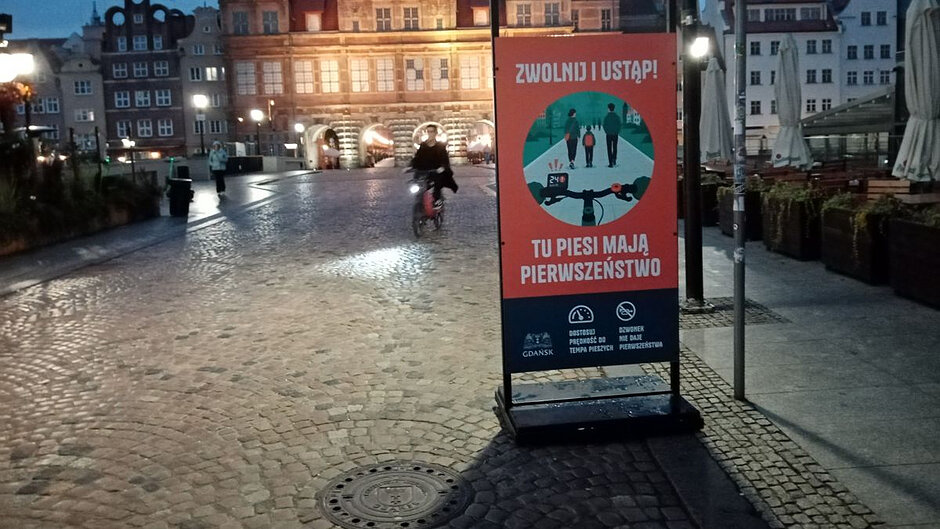 Na Moście Stągiewnym stoi ekspozytor w ramach akcji "Ustąp pieszym". W tle jedzie rowerzysta. 