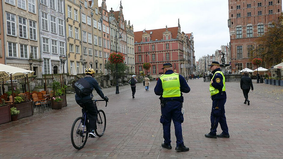 Na ulicy Długi Targ stoi dwóch strażników miejskich, a przejeżdza rowerzysta. W tle kilkoro przechodniów. 
