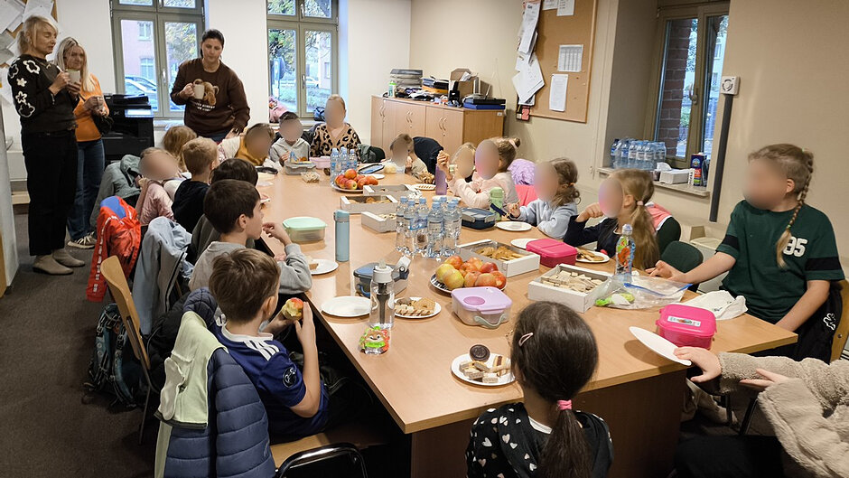 Na zdjęciach widoczna jest grupa dzieci uczestniczących w spotkaniu edukacyjnym w pomieszczeniu biurowym. Uczestnicy siedzą przy dużym prostokątnym stole, na którym znajdują się talerzyki z przekąskami, pudełka ze słodkimi wypiekami, owoce oraz liczne butelki z wodą. Dzieci rozmawiają i spożywają przygotowany poczęstunek. Wokół stołu ustawione są krzesła, a na ich oparciach wiszą kurtki i plecaki.

W tle znajdują się osoby dorosłe opiekujące się grupą. Pomieszczenie jest jasne, wyposażone w biurka, szafki, tablicę informacyjną oraz duże okna, przez które wpada światło dzienne. Atmosfera przypomina przerwę podczas zajęć lub spotkania integracyjnego.
