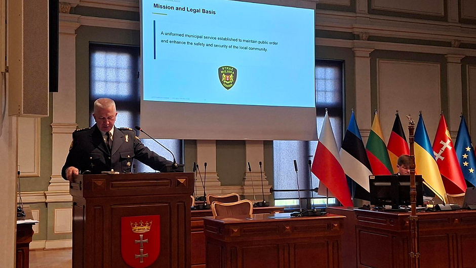 Komendant SMG Leszek Walczak przemawia podczas międzynarodowego seminarium. Za nim wyświetlona plansza prezentacji, z boku flagi państw europejskich.