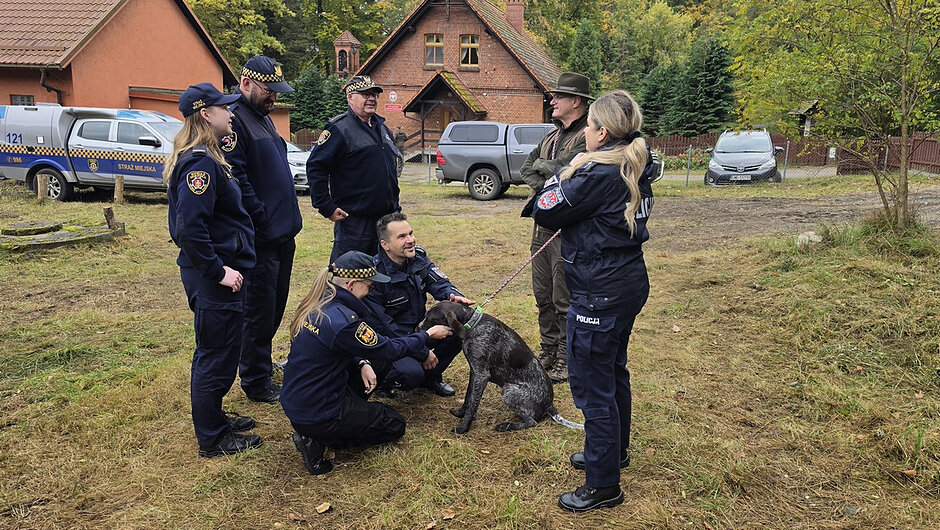 Funkcjonariusze Straży Miejskiej, Policji oraz przedstawiciele innych służb i instytucji spotkali się na terenie leśnym, aby wspólnie uczestniczyć w działaniach na rzecz ochrony środowiska. W centralnej części zdjęcia widoczny jest pies, wokół którego zgromadzili się uczestnicy wydarzenia. W tle znajdują się pojazdy służbowe oraz zabudowania położone na skraju lasu.