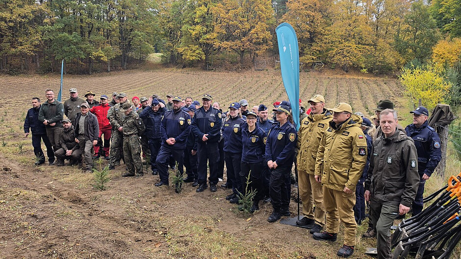 Uczestnicy akcji ustawili się do wspólnego zdjęcia na terenie przygotowanym pod nowe nasadzenia. W wydarzeniu biorą udział przedstawiciele różnych służb mundurowych, leśnicy oraz osoby zaangażowane w działania proekologiczne. W tle widoczne są młode sadzonki drzew oraz jesienny las.