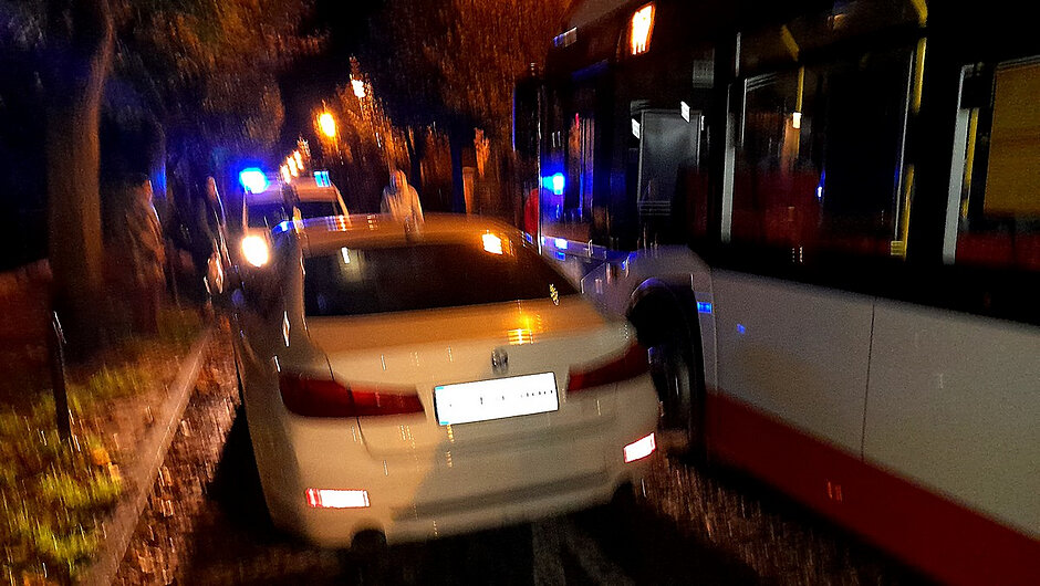 
Na zdjęciu widoczny jest biały samochód osobowy zaparkowany przy krawędzi jezdni. W tle, przy oświetlonej ulicy, widoczne są służby prowadzące czynności na miejscu zdarzenia. Fotografia została wykonana po zmroku, a sztuczne oświetlenie oraz sygnały świetlne pojazdów służbowych podkreślają interwencyjny charakter sytuacji. 