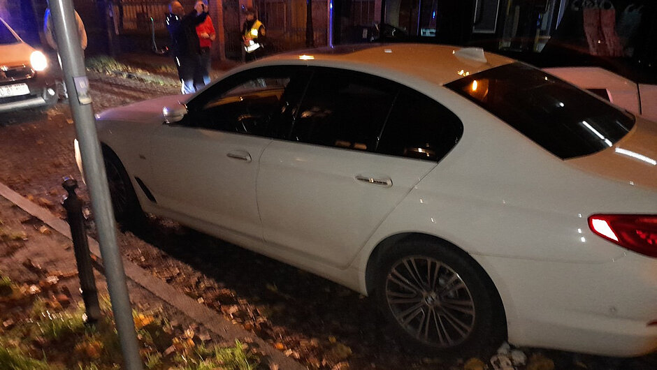 Drugie zdjęcie przedstawia skutki nieprawidłowego parkowania pojazdu w bezpośrednim sąsiedztwie torowiska tramwajowego. Biały samochód stoi tak blisko torów, że przejeżdżający tramwaj musiał zatrzymać się tuż obok pojazdu. W tle widoczne są pojazdy służb z włączonymi sygnałami świetlnymi oraz osoby uczestniczące w działaniach na miejscu. Fotografia obrazuje, jak jedno niewłaściwie zaparkowane auto może doprowadzić do utrudnień w ruchu komunikacji miejskiej i wpłynąć na podróż pasażerów.