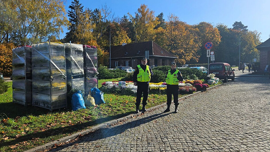 Dwójka strażników stoi na poboczu drogi przed Cmentarzem Srebrzysko. Obok stoją palety z kwiatami nagrobnymi. 