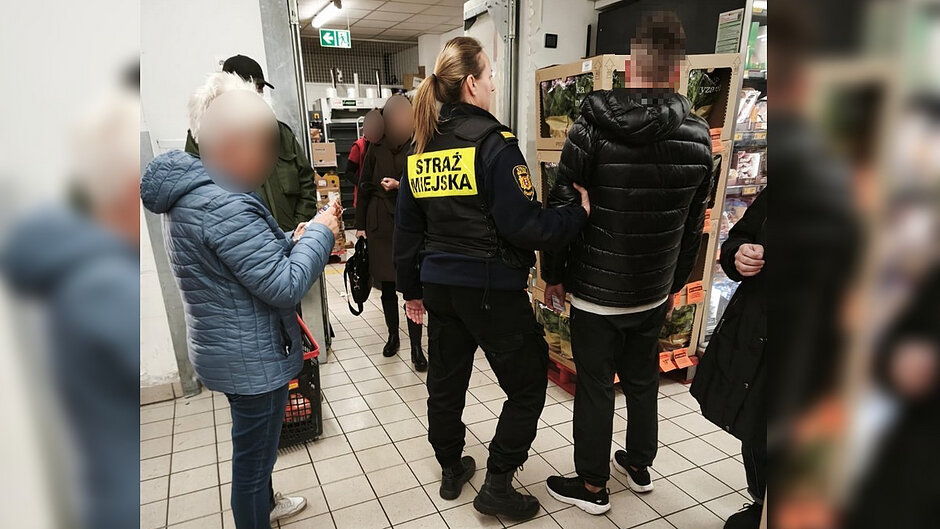 W sklepie spożywczym, na jasnej kafelkowej posadzce, stoi kilka osób. W centrum kadru znajduje się funkcjonariuszka Straży Miejskiej zwrócona tyłem do obiektywu. Na plecach ma żółto-czarną naszywkę z napisem „Straż Miejska”. Obok stoi osoba w czarnej kurtce, z którą funkcjonariuszka prowadzi czynności. Po lewej stronie widoczna jest osoba w niebieskiej kurtce trzymająca telefon. Twarze kilku osób zostały celowo rozmyte. W tle znajdują się sklepowe regały z towarem oraz oznaczenie wyjścia ewakuacyjnego. Zdjęcie przedstawia interwencję prowadzoną wewnątrz sklepu.