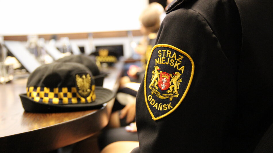 fragment rękawa z emblematem straży miejskiej w Gdańsku, w tle czapka strażnika leżąca na stole