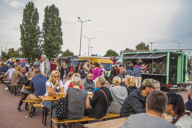 Zdjęcie przedstawia kolorowy tłum ludzi na placu zajadających różne potrawy. W tle widoczne stoisko food trucka