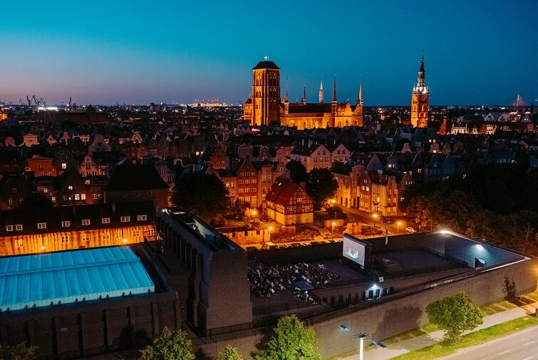 Zdjęcie przedstawia nocny Gdańsk. Na pierwszym planie wiele osób zasiada na dachu teatru szekspirowskiego. W tle rozświetlone główne miasto.